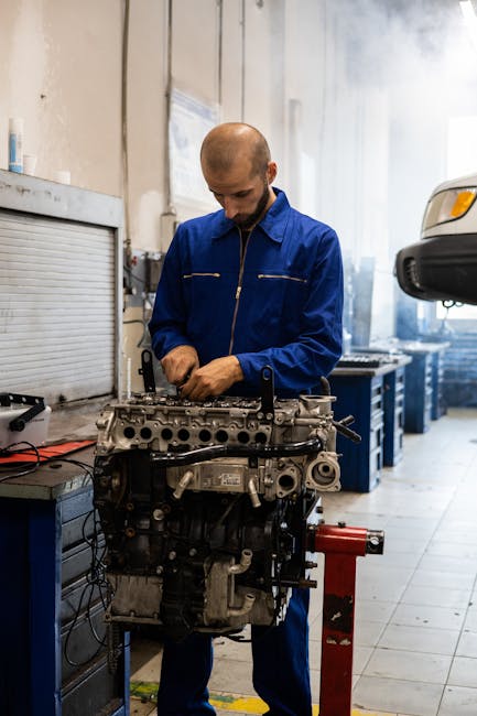 Mechanic in a blue uniform repairing an engine in a garage workshop.