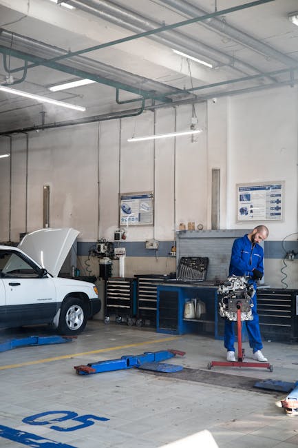 Mechanic in blue coverall working on car engine in a well-lit auto repair shop.